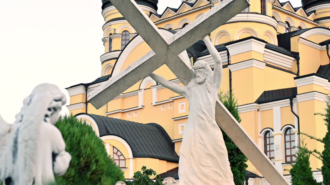 A statue of Jesus with a cross stands prominently in a garden, with a colorful church in the background. The sun shines down, highlighting the peaceful scenery
