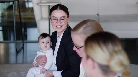 Una-Chica-Morena-Segura-De-Sí-Misma-Con-Gafas-Redondas-Y-Ropa-De-Negocios-Sostiene-A-Un-Niño-Pequeño-En-Sus-Brazos-Durante-Su-Reunión-De-Negocios-En-La-Alfombra-Durante-Un-Descanso-Entre-El-Trabajo.