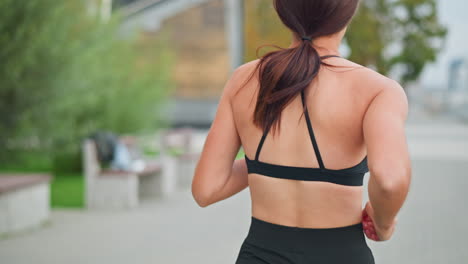 back view of lady in black neck top running along park path with blurred background showing trees and bench, promoting outdoor fitness and healthy lifestyle