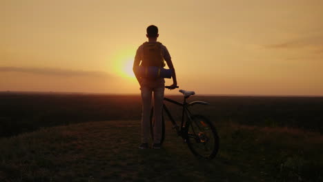 A-Man-With-A-Backpack-And-A-Bicycle-It-Stands-And-Looks-At-The-Horizon-Where-The-Sun-Sets-Cycling-An