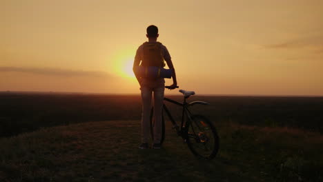 a man with a backpack and a bicycle it stands and looks at the horizon where the sun sets cycling an