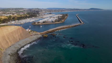 Stunning-aerial-view-of-sandstone-sea-cliff,-jetty,-waves,-harbor,-boats