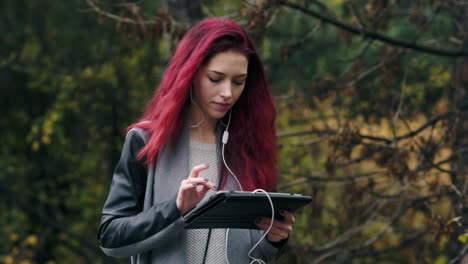 joven hermosa con el cabello rojo camina en el parque de otoño y escucha música a través de una tableta. joven mujer atractiva con una tableta digital en las manos