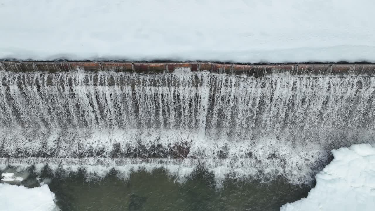 A drone moves from right to left, capturing the water cascading over the retaining boards of a dam during winter, with ice forming along the edges of the flowing water.