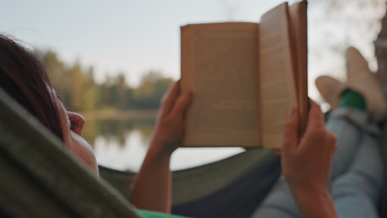 Donna che legge un libro su un'amaca in riva al lago, profilo rilassato con le gambe distese e il libro tenuto sopra il petto, luce solare soffusa sull'acqua, brezza leggera tra gli alberi, un'atmosfera da campeggio informale e pacifica.