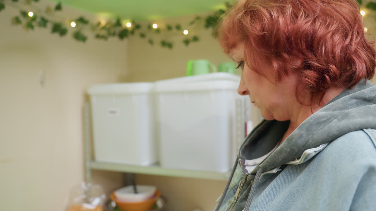 white woman reaches for container, pausing cozy kitchen storage scene with string lights, plastic bins on metal shelf, red hair visible, denim jacket and hoodie, slow deliberate movements, expressions