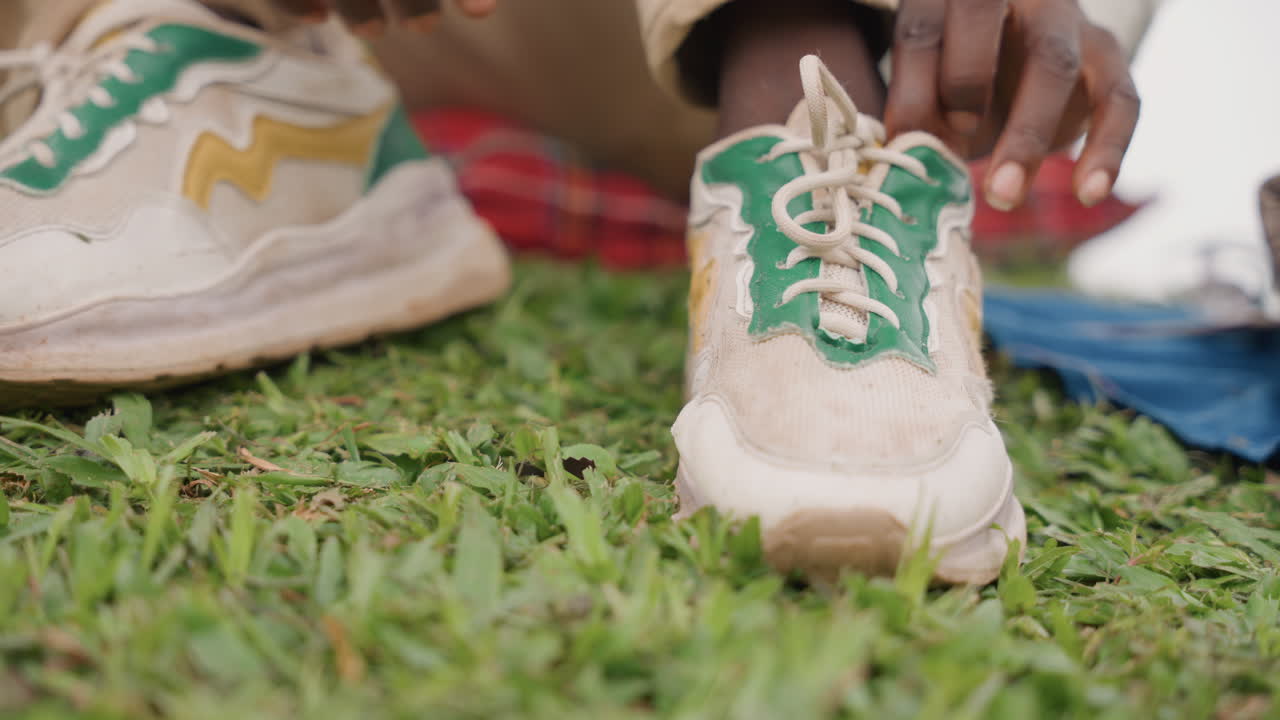 Padre di colore che insegna al figlio ad allacciare le scarpe sull'erba, primo piano di mani pazienti che annodano i lacci accanto a scarpe da ginnastica e coperta a quadri, preparazione sportiva prima dell'allenamento, istruzioni gentili al parco