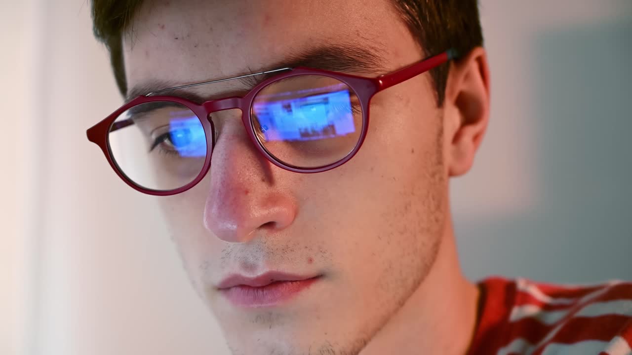 Young man in pink-striped sweater scrolling down a page. Reflection in his red glasses. Front view. Close up