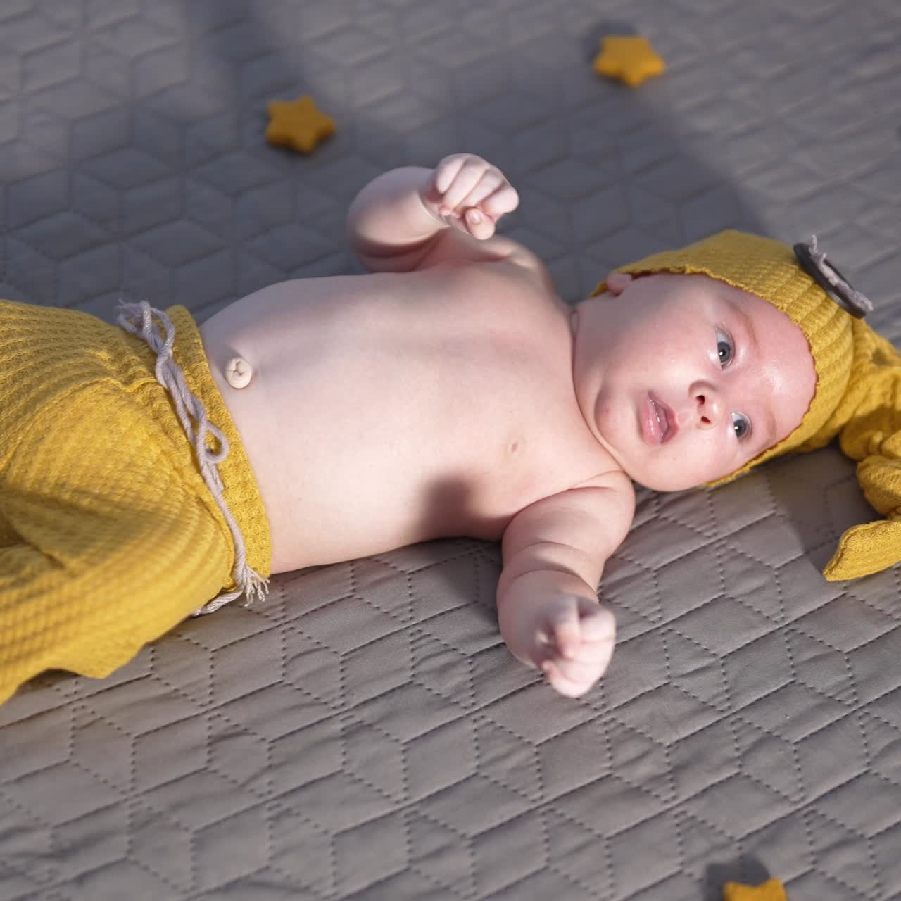 Adorable child in yellow hat and pants lying on the bed face up. Baby in yellow costume with decorative toy stars. View from above