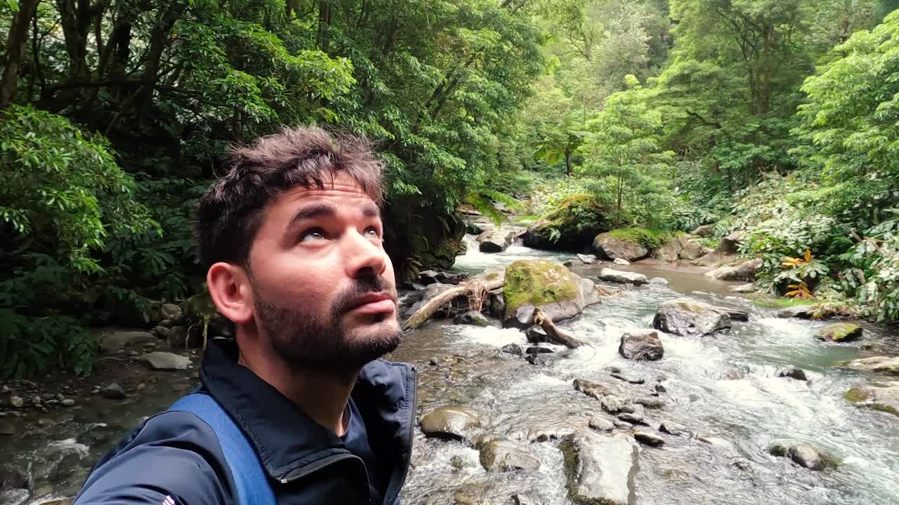 Selfie di un uomo che guarda la fotocamera, circondato da alberi e dall'acqua del fiume nel bosco.