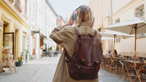 Vista-Trasera-De-Una-Joven-Turista-Con-Mochila-Escuchando-Música-A-Través-De-Auriculares-Caminando-Por-La-Calle