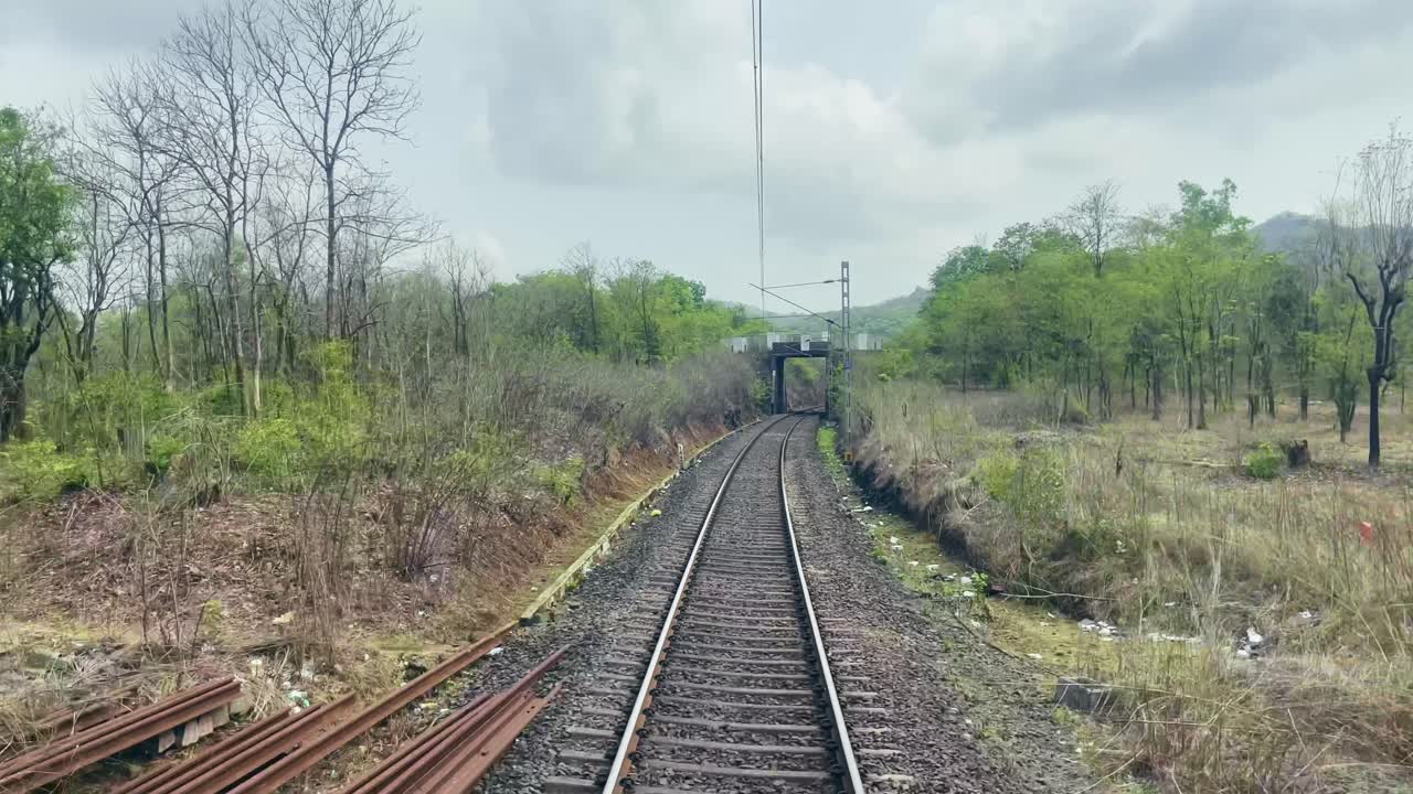 A railway track runs through gentle hills on both sides, with a small bridge arching overhead. The sun shines warmly, with fluffy white clouds Maharashtra India 4K
