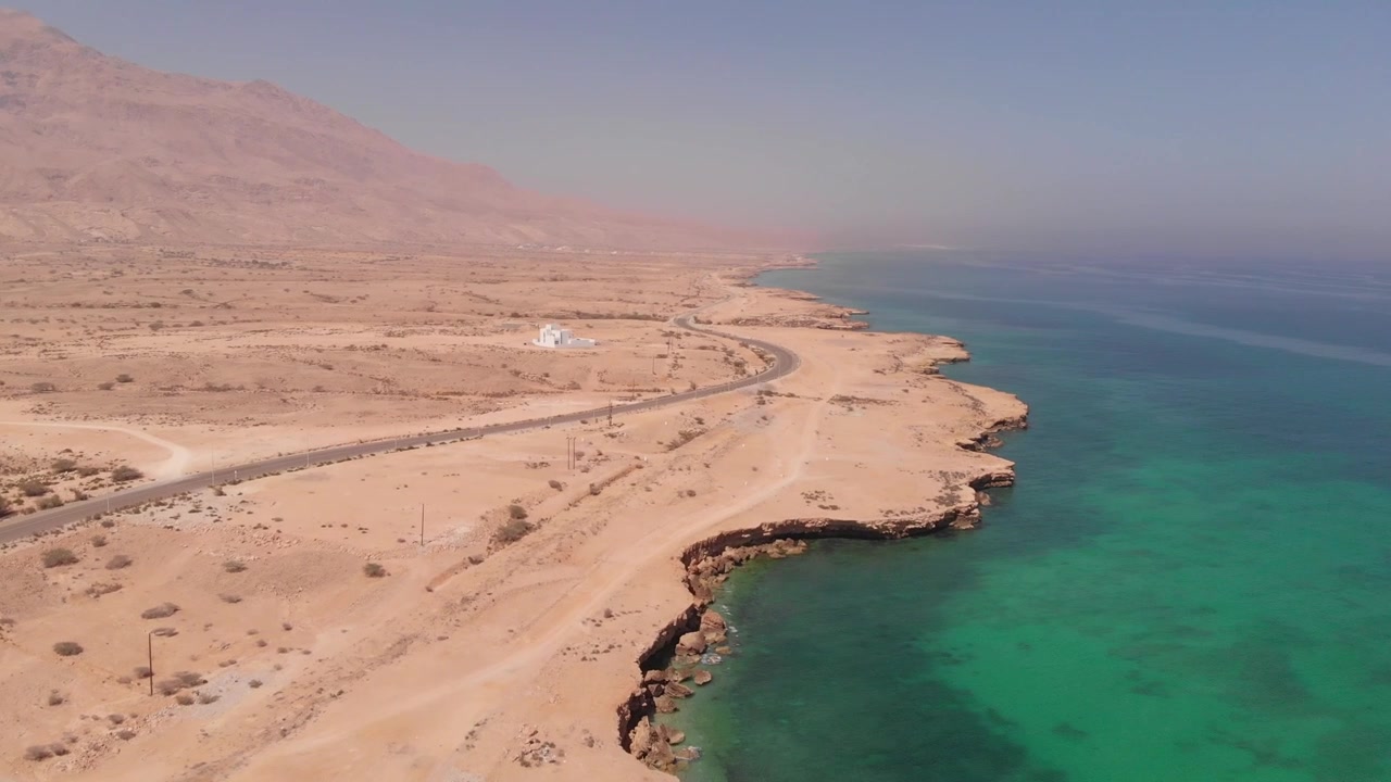 Ripresa aerea con drone dall'alto di una strada costiera tortuosa lungo le onde turchesi dell'oceano e le aspre scogliere desertiche dell'Oman. Mette in mostra il drammatico contrasto tra paesaggio arido e mare.