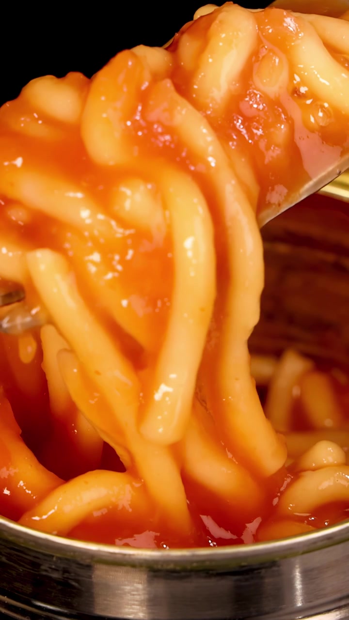 Una forchetta di metallo solleva spaghetti al sugo da una lattina aperta sotto un'illuminazione brillante e uniforme. La telecamera rimane ferma, concentrandosi sulla consistenza e sul movimento della pasta.