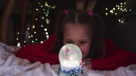 chica caucásica sonriendo y mirando el globo de nieve mientras yacía bajo la manta fuerte durante la navidad en