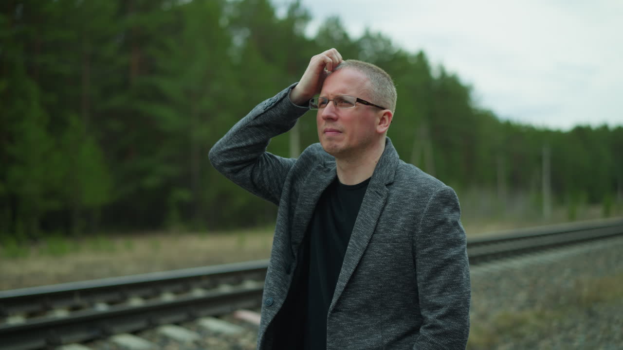 Close-up view of a man in glasses and a gray jacket standing on a railway track, looking worried as he rubs his head with one hand