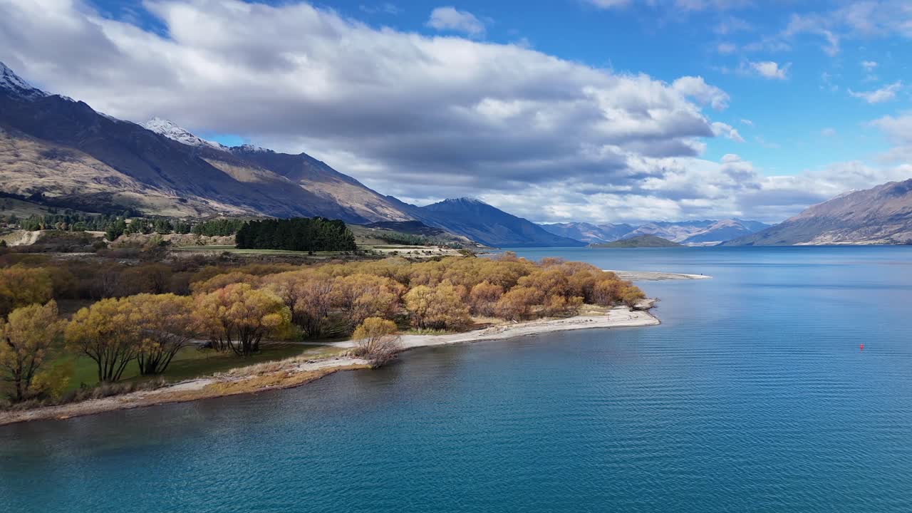 Aerial footage captures Glenorchy's tranquil lakeside with vibrant autumn foliage and clear blue waters under a partly cloudy sky