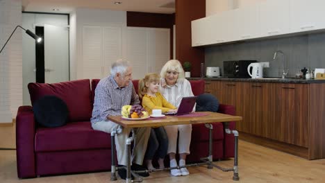 abuelo y abuela sentados en la sala de estar y enseñando a la nieta pequeña usando una computadora portátil