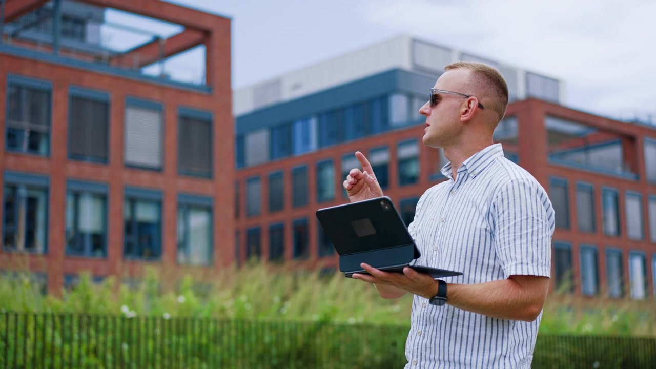 Cheerful man holding closed tablet standing near modern brick office building. Smiling male in striped shirt and dark sunglasses poses outdoors by green vegetation and urban corporate architecture.