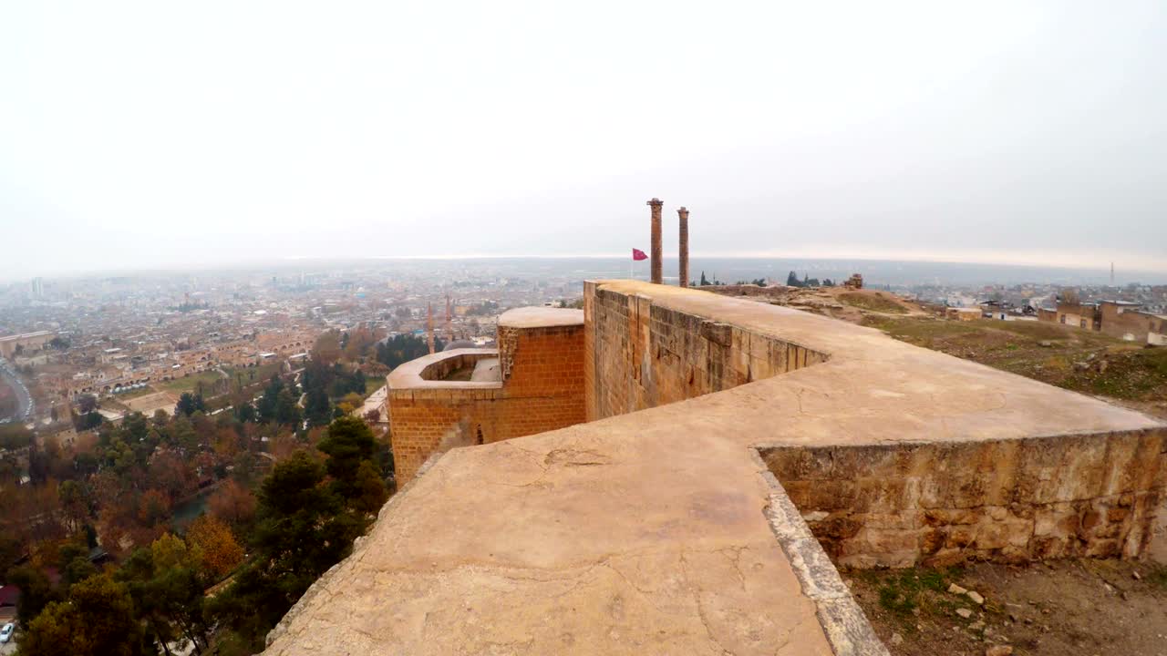 Muro di cinta del Castello di Urfa con sullo sfondo la città a due colonne e la bandiera rossa turca al vento, sotto neve e pioggia.