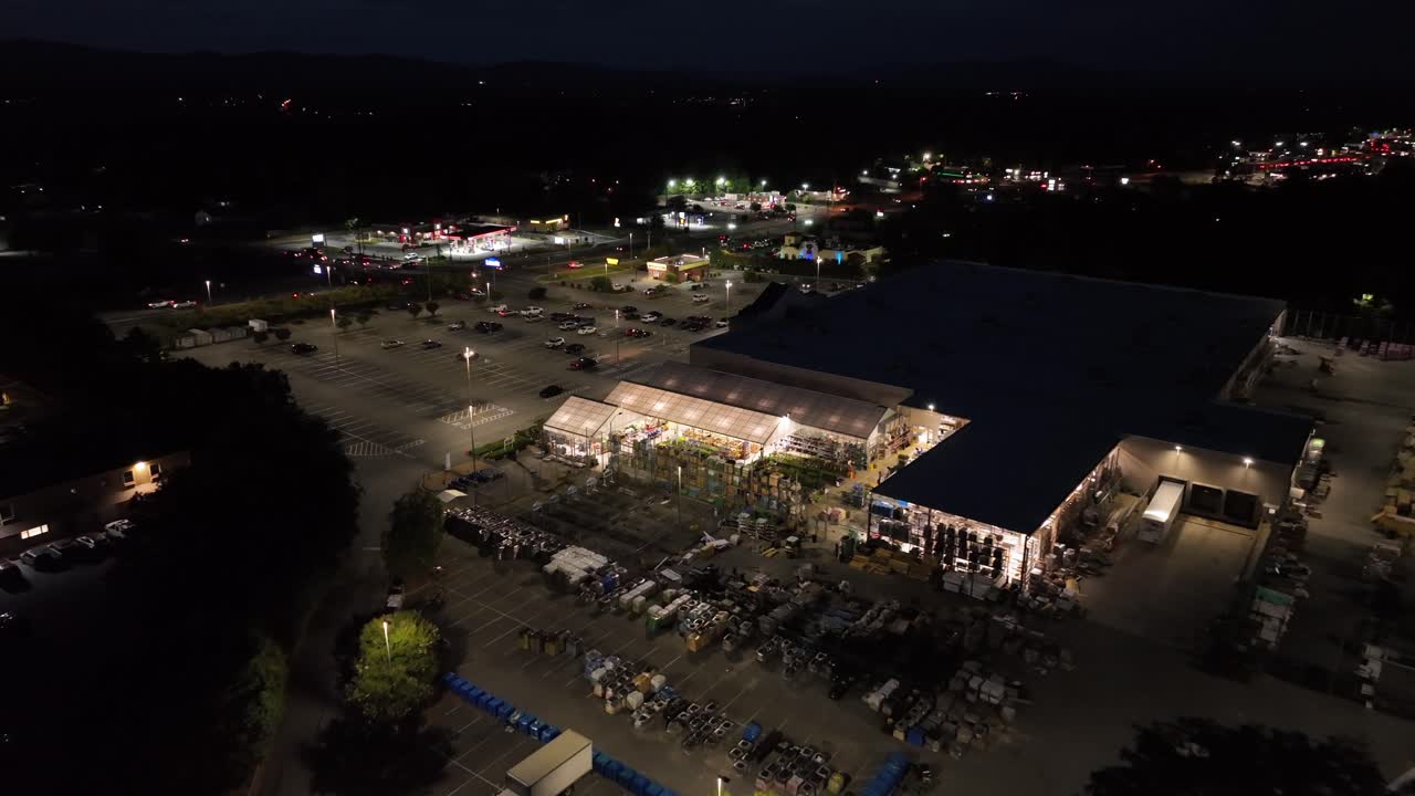 Lighting shopping center with plants at night. Aerial flyover. Parking area. American city with traffic in suburb of Lynchburg, Virginia. Top down. Home improvement garden center