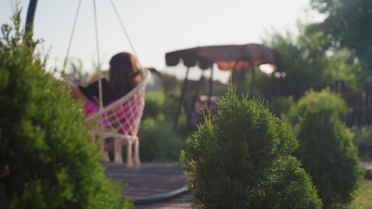 Woman Enjoying Sunny Outdoor Lounge, Serene Woman Basking In Afternoon Sunlight Sitting Comfortably, Relaxed Woman Seated Outdoors In Hanging Chair Amid Lush Foliage During Warm Sunlight