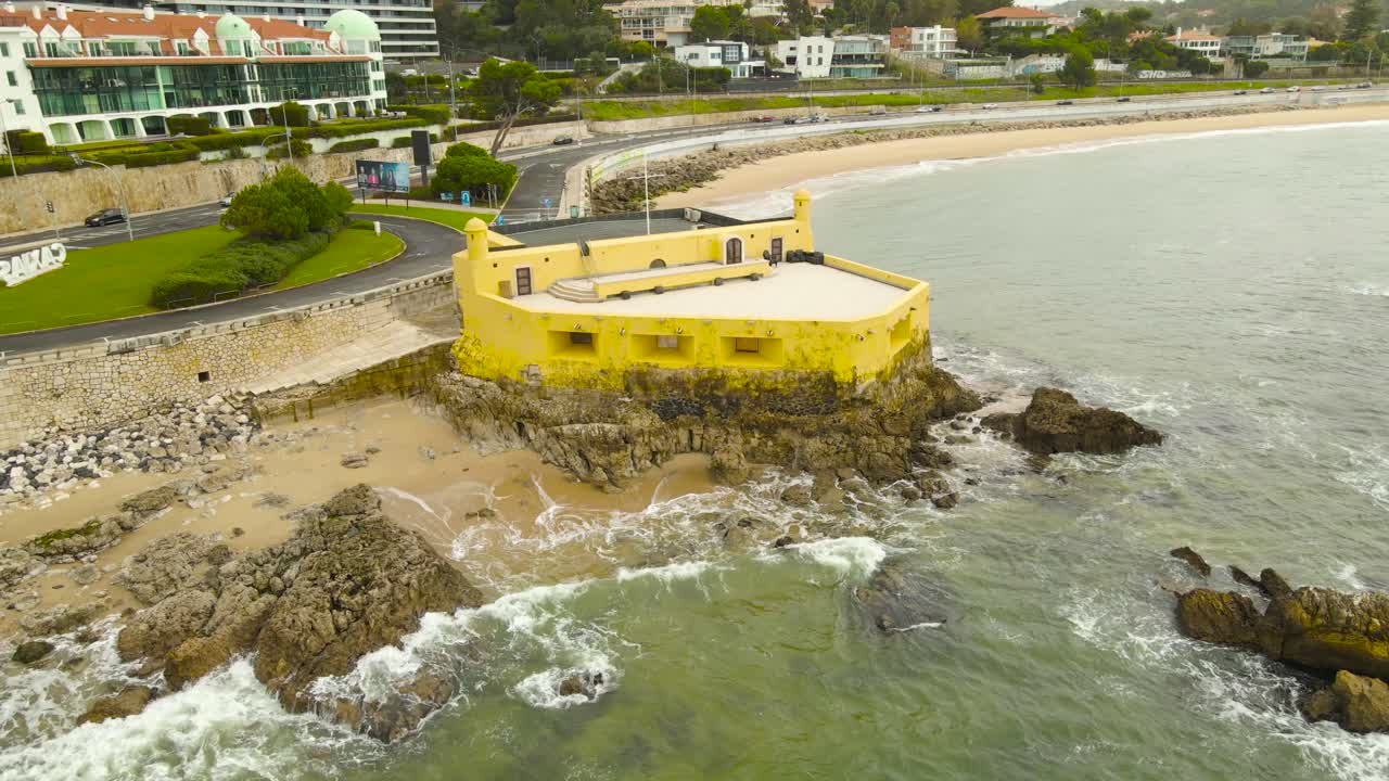 Aerial drone footage of Forte da Giribita and rocky coastline in Caxias Portugal with ocean waves hitting stone reefs beside a coastal highway and seaside promenade on a cloudy day