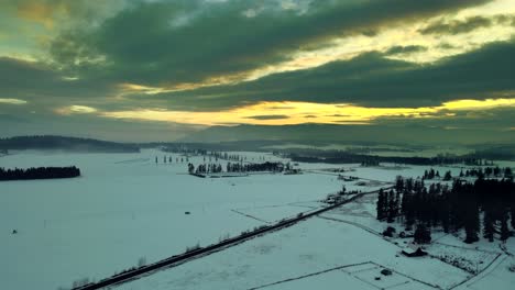 Toma-Aérea-De-Tierras-De-Cultivo-Bajo-Un-Impresionante-Cielo-Amarillo-Rodeado-De-Pastizales-Cubiertos-De-Nieve-Y-Bosques-De-Agujas