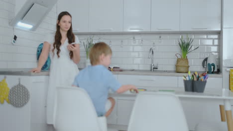 Mamá-En-La-Cocina-Lavando-Platos-Y-Dos-Hijos-Sentados-En-Un-Escritorio-Dibujando-En-Papel-Con-Lápices-De-Colores.-La-Joven-Madre-De-Familia-Sonríe-Y-Mira-A-Los-Niños.