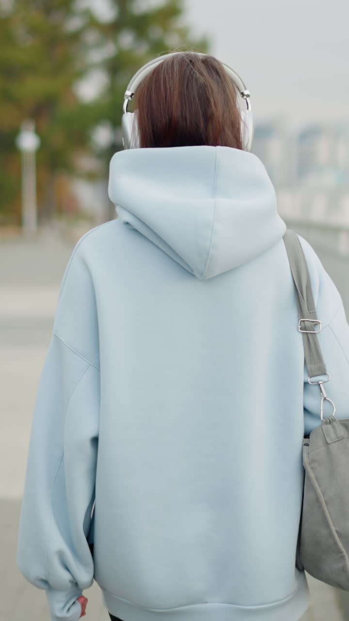 Back view of woman in blue sweater and headphones holding bag, walking slowly along iron rail, serene outdoor atmosphere, peaceful morning walk, urban setting, casual lifestyle