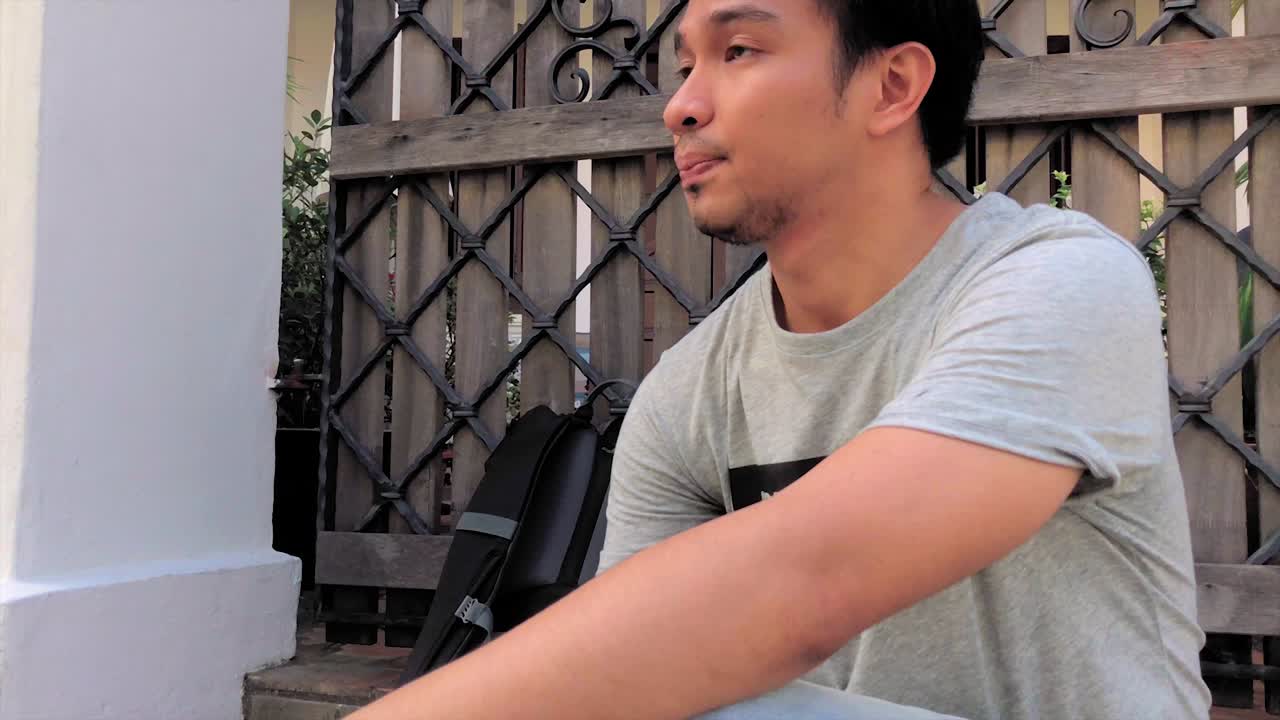 Tired Young Millennial Man Slowly puts down Backpack and sits in front of a Wooden Gate Door at Daytime. Slow Motion and Follow Shot
