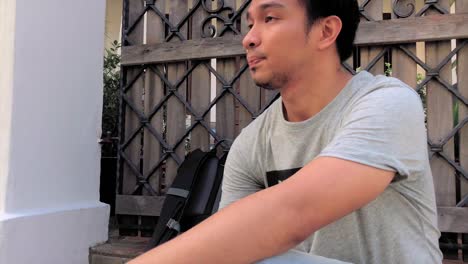 tired young millennial man slowly puts down backpack and sits in front of a wooden gate door at daytime. slow motion and follow shot