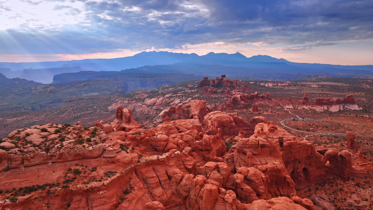 Sorvolo delle cime arrotondate delle rocce rosse nell'arido deserto. I raggi del sole al tramonto attraversano il cielo nuvoloso grigio. L'Arches National Park nello Utah, USA