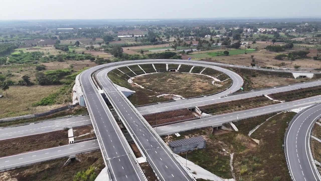 Drone shot capturing the Chennai–Bengaluru Expressway showcasing structured highway engineering, wide lanes, interconnected flyovers, and geometric road patterns stretching across rural terrain.