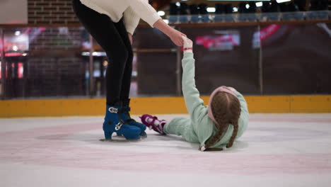 a close view of a mother helping her daughter up from the ice rink, the child is lying on the ground in a mint green outfit with pink skates is gently pulling her up