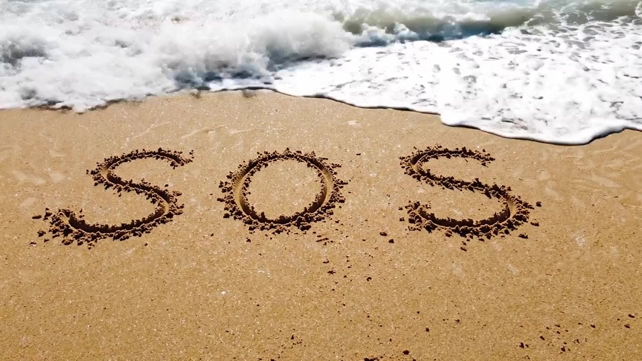 Una disperata richiesta d'aiuto: il messaggio SOS scritto nella sabbia sulla riva mentre le onde lo lambiscono, simboleggiando urgenza e angoscia in riva all'oceano.