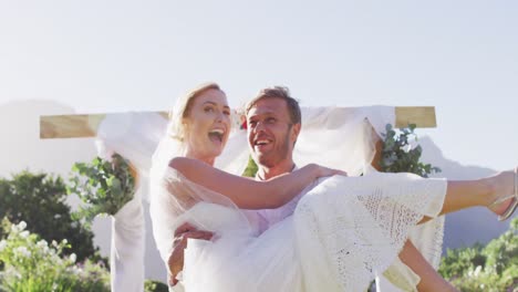 Feliz-Marido-Caucásico-Lleva-A-La-Novia-Recién-Casada-Y-Camina-Por-El-Altar-Al-Aire-Libre