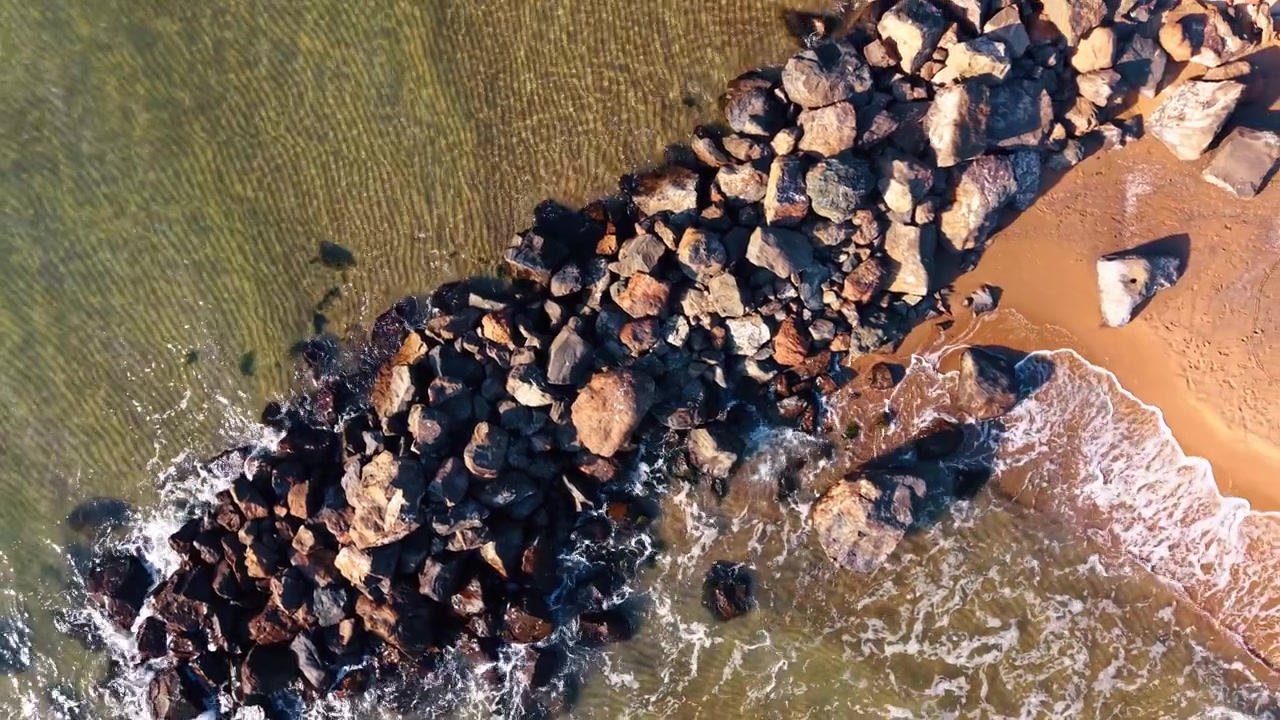 Vista dall'alto di un frangiflutti roccioso tra acque calme e turbolente. Prospettiva drone dall'alto di grandi massi che dividono secche dorate poco profonde e la risacca bianca