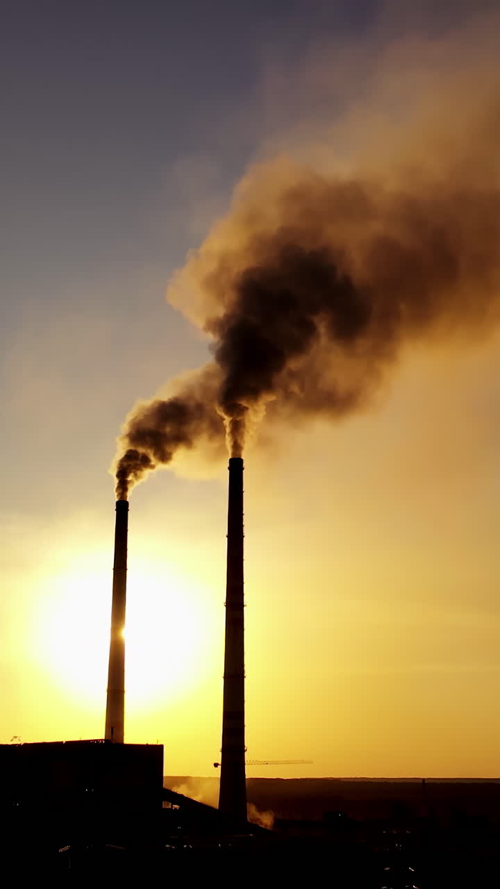 Dark silhouette of a factory at sunset. Pipes with dirty smoke of metallurgical plant in the evening. Vertical video