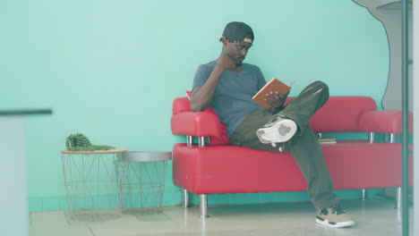 relaxed man in casual shirt and cap sits on red couch with crossed leg, holding open book and reading intently with hand resting on face, focused on literature in bright indoor setting