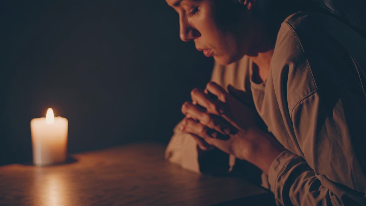 A dimly lit video scene shows a person in soft focus, hands clasped on a table beside a candle