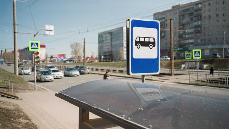 close view of city traffic on a sunny day with bus stop and pedestrian signs in the foreground, high-rise buildings, and people walking, cars waiting for them to cross the road