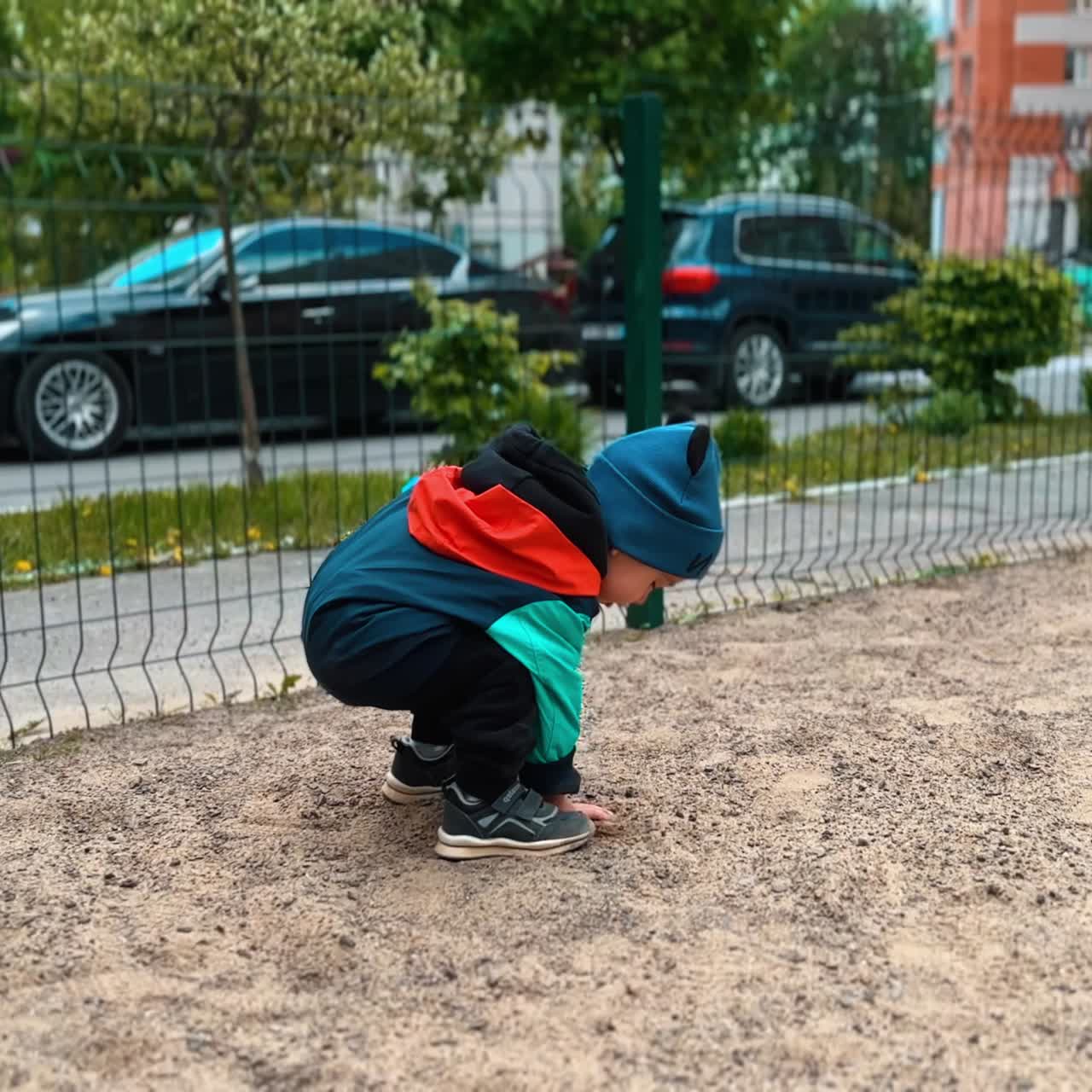 Little kid picks up some sand from the ground and throws it away through the fence. Toddler has active time outdoors