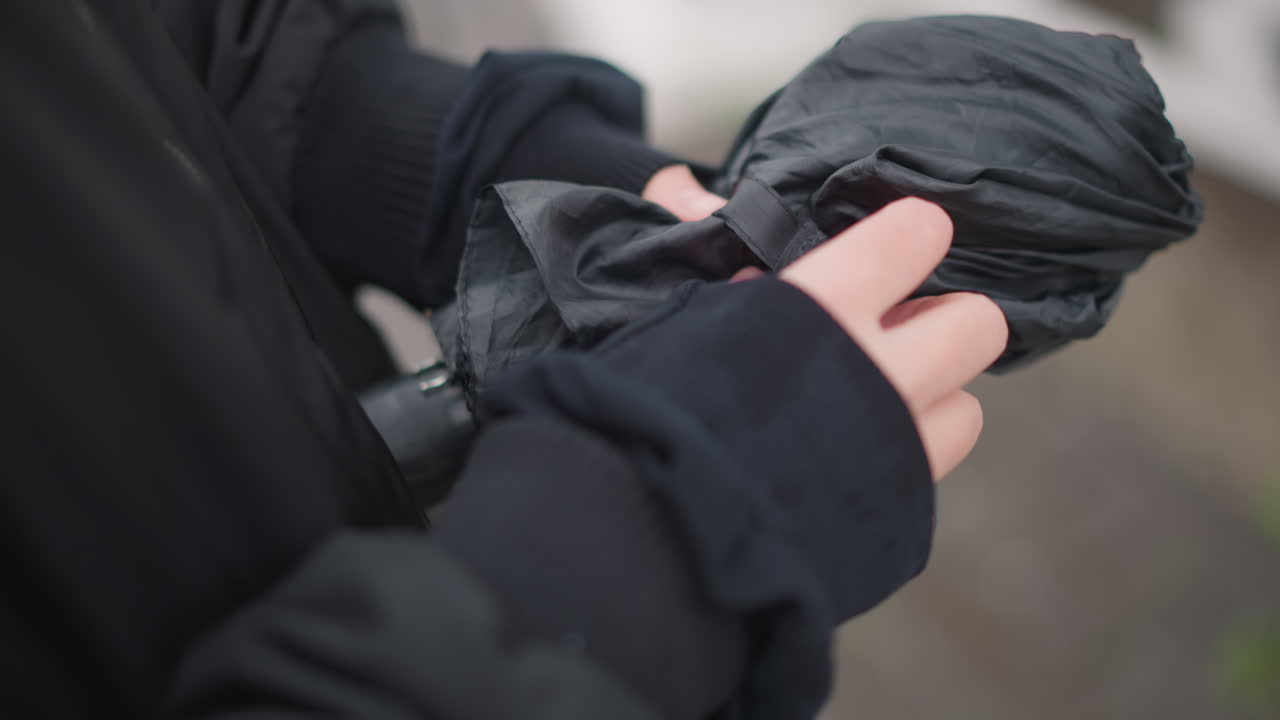 Woman securing umbrella strap with relieved motion, closeup of hands fastening folded canopy, textured nylon and droplets, muted city scene and sidewalk reflections, pedestrian finishing commute