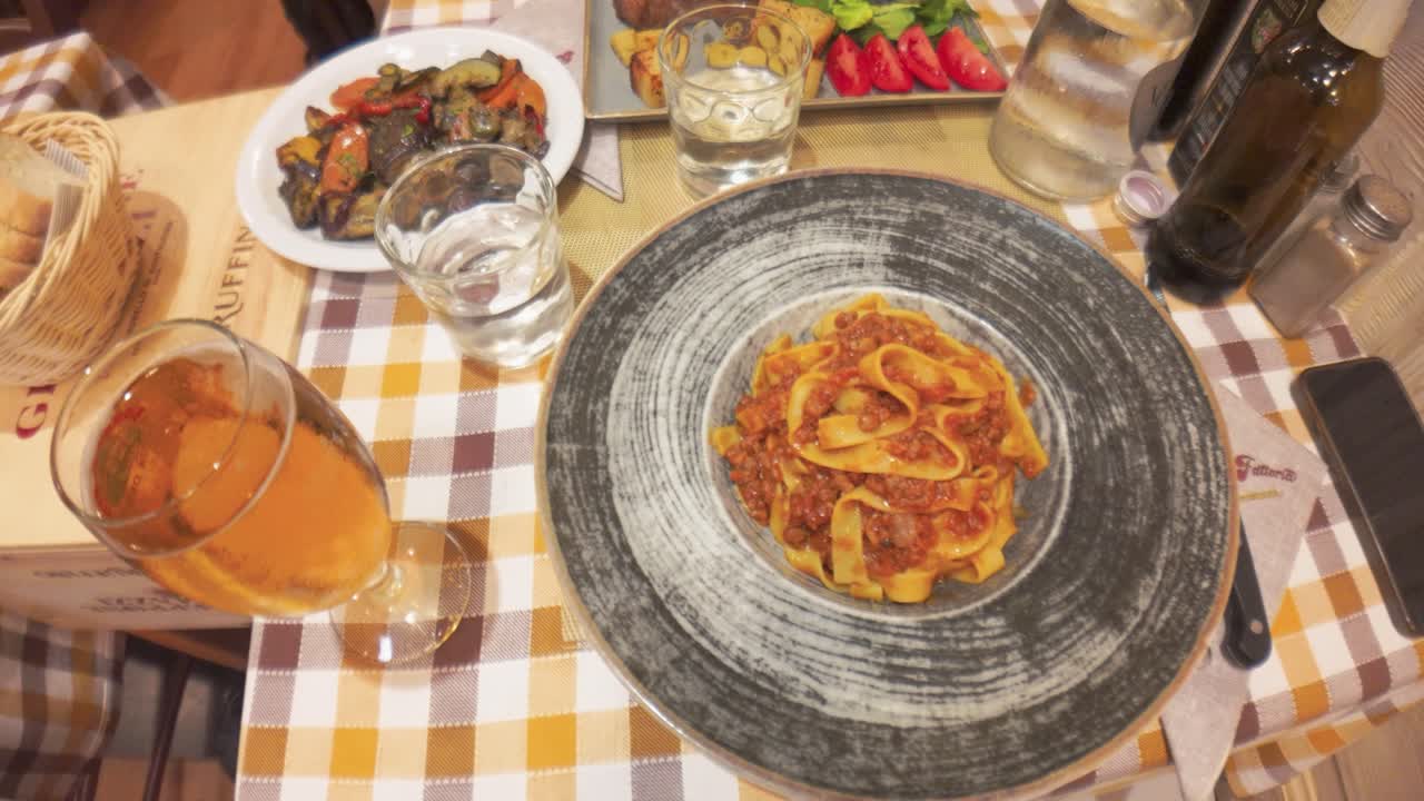 Statische Aufnahme von oben von rustikaler italienischer Pasta, gegrilltem Gemüse und Getränken, arrangiert auf einem karierten Tischtuch in warmer, einladender Restaurantbeleuchtung. Keine Personen sichtbar für saubere Stocknutzung.