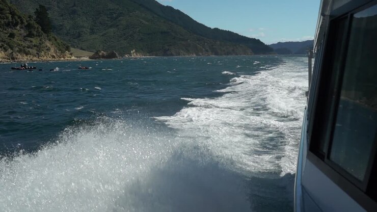 Onde spumeggianti che si infrangono sul fianco di una nave da crociera nei Marlborough Sounds, Nuova Zelanda, con colline verdi sullo sfondo