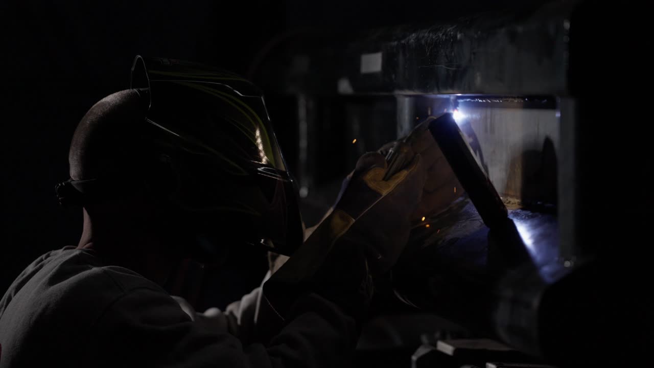 Scintille che volano al rallentatore mentre un saldatore mascherato utilizza una torcia per riparare un veicolo da costruzione in un cantiere