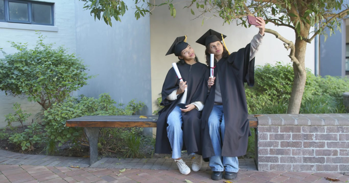 Laureate afroamericane in toga con diplomi che aprono il telefono e posano per un selfie di laurea su una panchina.