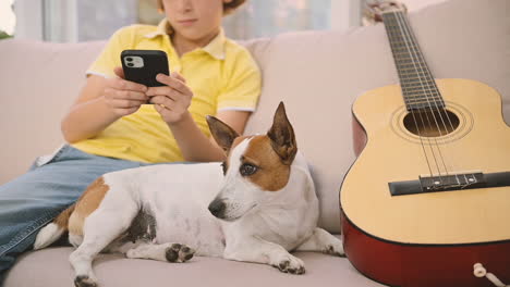 blond boy sitting on the sofa using a smartphone, next to his dog lying near a guitar