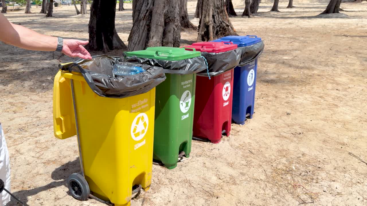 A hand drops a plastic bottle into a yellow recycling bin on sandy ground